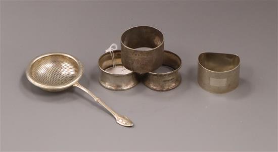 Four silver serviette rings and a silver tea strainer.